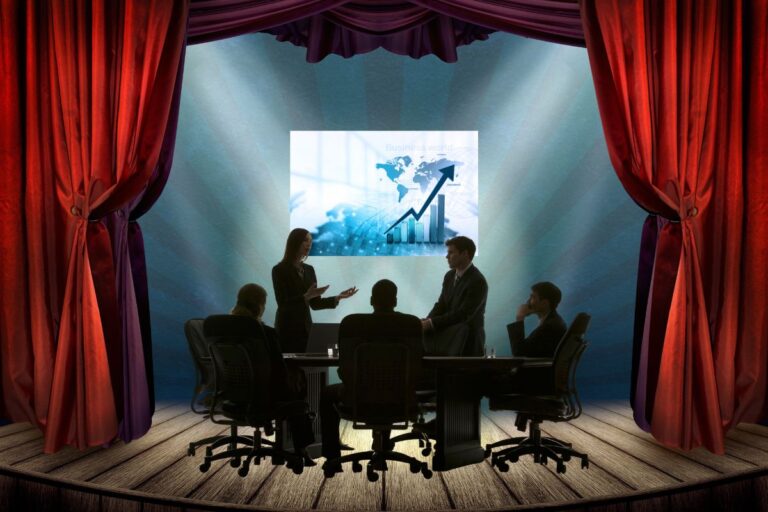 Business people on a theater stage, sitting at a table looking at a graph on the wall.